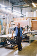 © Maskot - Full length of smiling male carpenter standing with hand on hip by workbench