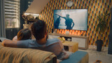 Fototapeta  - Couple sitting and hugging on sofa in cozy living room, watching action movie on TV or criminal blockbuster on streaming service, resting at home after workday. Home theater in modern apartment.