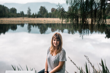 Canvas Print - blonde caucasian girl in gray t-shirt and black pants looking at camera sitting on the shore of clear water lake near forest and mountains cloudy gloomy day, new zealand glaciers