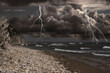 Dark cloudy sky with lightnings over sandy beach with rocks and sea. Thunderstorm