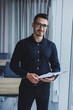 © Дмитрий Ткачук - A Caucasian business man in glasses and a black shirt sits in the office and looks at sales growth charts. A young successful businessman in classic clothes stands by the window in the office