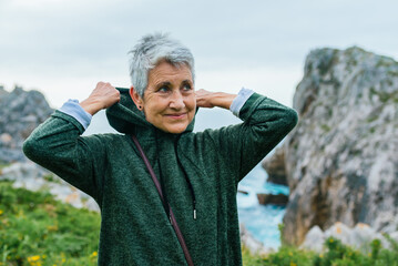 older woman with gray hair, wearing sportswear, pulling up the hood of her sweatshirt as she jogs along a coastal path. sports activities in the elderly.