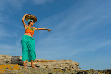 A woman in boho style clothes with a shamanic ritual tambourine on the rocks on the seashore on a clear sunny day. The concept of freedom of mind and body in nature