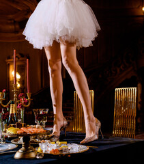 Cropped of elegant legs of woman wearing on short puffy dress and snakeskin high heels, standing on celebrating table with dishes and gold chairs around, placed on luxury aristocratic palace