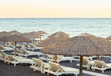 White Color Sunlounger Chairs On Black Sand Beach With Natural Reed Sun Umbrellas At Sunset. Holiday In Greece Concept, Lot Of Copy Space.