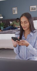 Wall Mural - vertical video of biracial woman in bedroom using smartphone and smiling, partner in background