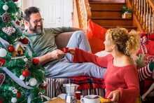Happy young adult couple at home enjoying christmas morning in december taking coffee and breakfast together. Man smile to a woman in holiday leisure activity indoor sitting on the sofa. Xmas tree