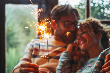 Love and romantic leisure activity with man and woman holding fire sparkler light together hugging and enjoying relationship at home sitting on a sofa. Christmas time for happy couple alone indoor