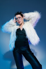 Wall Mural - model in black corset and trendy faux fur jacket posing with hands behind head on blue background.