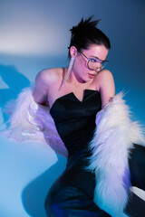 Wall Mural - brunette model in black corset and white faux fur jacket sitting on blue background.