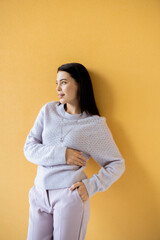 Wall Mural - smiling brunette woman in knitted sweater posing with hand in pocket and looking away on yellow background.