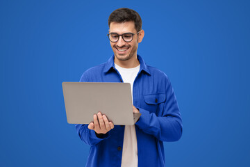 Happy man standing with opened laptop, browsing online or typing message, isolated on blue