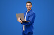 © Damir Khabirov - Young man standing holding laptop and looking at camera with happy smile, isolated on blue