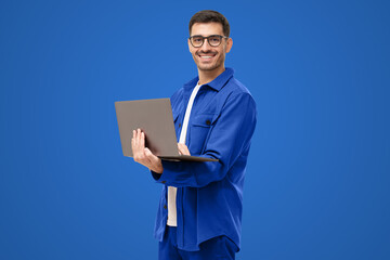 Young man standing holding laptop and looking at camera with happy smile, isolated on blue