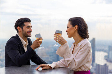 Poster - Caucasian couple celebrating at luxury skyscraper rooftop bar at summer sunset. Confident man and woman couple enjoy city lifestyle having dinner and cocktail drink at outdoor restaurant in evening.