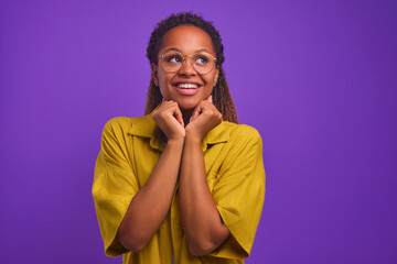 Wall Mural - Young beautiful long haired African American woman raises hands to chin and presses to chest in anticipation of long-awaited meeting or presenting gift stands on purple studio background