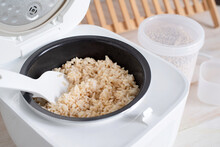 Opened electric rice cooker with cooked steaming brown rice on wooden counter-top in the kitchen