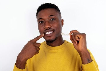 Wall Mural - young handsome man wearing yellow sweater over white background holding an invisible aligner and pointing to her perfect straight teeth. Dental healthcare and confidence concept.