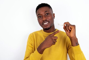 young handsome man wearing yellow sweater over white background holding an invisible aligner and pointing at it. Dental healthcare and confidence concept.