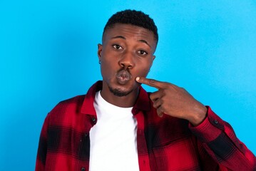 Wall Mural - Charming young handsome man wearing overshirt over blue background pointing on pout lips with forefinger, showing effect after lifting procedure,