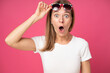 impressed shocked young attractive woman dressed in white t-shirt and holding sunglasses on her head with open mouth omg and big eyes isolated over pink red background