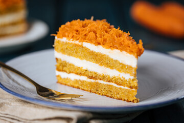 Wall Mural - Carrot cake with cream cheese in layers and carrots on top on a  blue and white background with carrots in blurry background