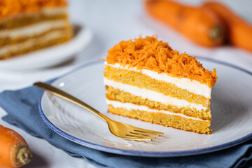 Wall Mural - Carrot cake with cream cheese in layers and carrots on top on a  blue and white background with carrots in blurry background