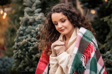 A Young Curly Woman Wrapped In A Warm Plaid In A Cell Sits Waiting For The New Year. The Girl On The Background Of Green Christmas Trees On The Street. Holiday Mood