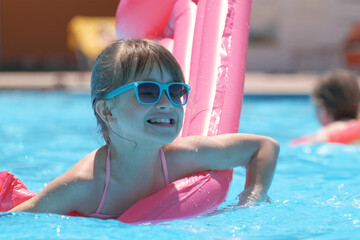 Young joyful child girl having fun swimming on inflatable air mattress in swimming pool with blue water on warm summer day on tropical vacations. Summertime activities concept