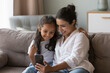 © fizkes - Attractive Indian woman and little cute daughter sit on sofa look at smartphone screen enjoy new mobile apps, calling through video call to family, spend time at home use cellphone. Modern tech, fun