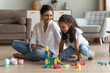 © fizkes - Indian woman and daughter play wooden blocks seated on floor smiling looking happy, enjoy carefree weekend leisure and pastime, having fun together at modern living room. Playtime at home with child