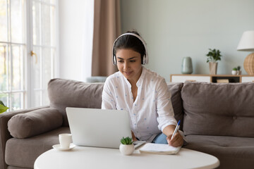 Canvas Print - Indian woman wear headphones holds pen making notes, writing assignment, doing exercise, practice foreign language, e-learning at home using laptop. Video call application usage for distance studying