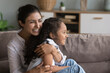 © fizkes - Loving laughing Indian mother hugging her daughter tightly seated on sofa enjoy tender moments spend priceless time together feeling unity and love. Caring parent and children bonding and protection