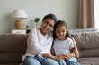 © fizkes - Portrait of happy Indian woman hugs little adorable daughter laughing seated together on sofa staring at camera enjoy pleasant communication through video conference app. Family bond, motherhood, fun