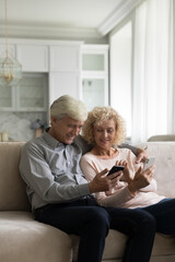 Vertical shot, aged couple sit on couch hold two smartphones, enjoy new gadgets, learn mobile application, help each other with device. Older generation use modern tech for comfort life, apps usage