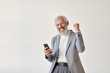 Excited overjoyed happy old senior business man investor winner raising fist holding cell using smartphone winning mobile bet money prize holding cellular phone celebrating victory isolated on white.