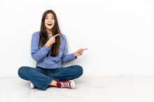 Young Woman Sitting On The Floor Surprised And Pointing Side