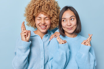 Wall Mural - Luck and expectations concept. Happy multiethnic women cross fingers make wish have desire dreams come true dressed in knitted jumpers stand closely to each other isolated over blue background.
