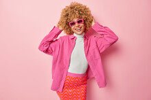 Indoor Shot Of Cute Curly Haired Woman Wears Sunglasses Shirt And Skirt Keeps Hands On Head Smiles Gladfully Feels Happy To Buy New Clothes Isolated Over Pink Background. People And Style Concept
