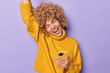 © wayhome.studio  - Overjoyed curly haired European woman exclaims loudly keeps arm raised up dressed in casual yellow jumper celebrates great news holds mobile phone gets message from boyfriend isolated over purple wall