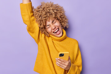 Wall Mural - Overjoyed curly haired European woman exclaims loudly keeps arm raised up dressed in casual yellow jumper celebrates great news holds mobile phone gets message from boyfriend isolated over purple wall