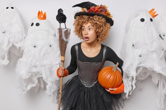 Magic and miracles concept. Impressed curly haired young woman witch poses with broom and pumpkin celebrates Halloween autumn holiday on 31st of October surrounded by spooky creatures. Scary day
