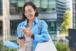 © wayhome.studio  - Outdoor shot of positive Asian woman holds cellular gadget and takeaway coffee checks received notification wears spectacles and blue shirt poses against urban background creats social publication