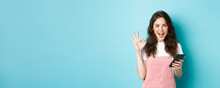 Image Of Excited And Pleased Young Woman Say Yes After Using Smartphone App, Online Shopping With Mobile Phone And Showing OK Sign, Standing Over Blue Background