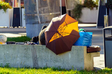 A Homeless Man Sleeps On A Bench Covered With An Umbrella Next To His Bags.