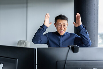 Wall Mural - Happy business broker looking at two paired computer monitors and happy, businessman working inside modern office building, asian man celebrating successful deal and signed contract.