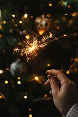 Wall Mural - Happy New Year! Burning sparkler in female hand on background of christmas tree lights in dark room. Atmospheric celebration. Hand holding firework against stylish decorated tree with illumination