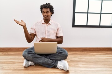 Sticker - African man with curly hair using laptop sitting on the floor smiling cheerful presenting and pointing with palm of hand looking at the camera.