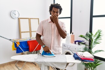 Wall Mural - African man with curly hair ironing clothes at home laughing and embarrassed giggle covering mouth with hands, gossip and scandal concept