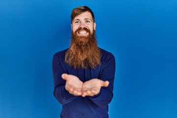 Poster - Redhead man with long beard wearing casual blue sweater over blue background smiling with hands palms together receiving or giving gesture. hold and protection